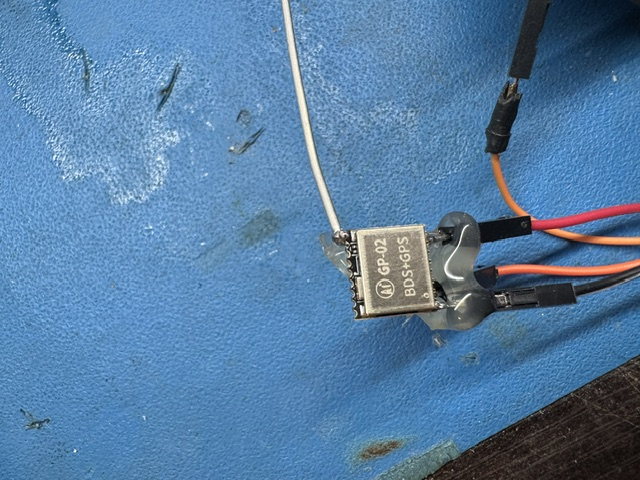 Close-up of a GNSS receiver module on the bench: a white antenna lead, taped wire joins and jumper wires running to a pin header.